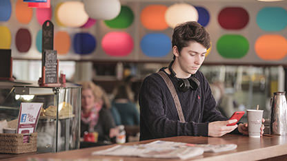 Teenager with headphones uses his phone whilst drinking a coffee.